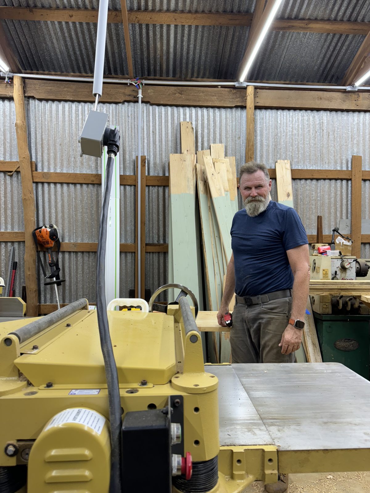 Standing at the thickness planer in the woodshop, lumber racked behind
